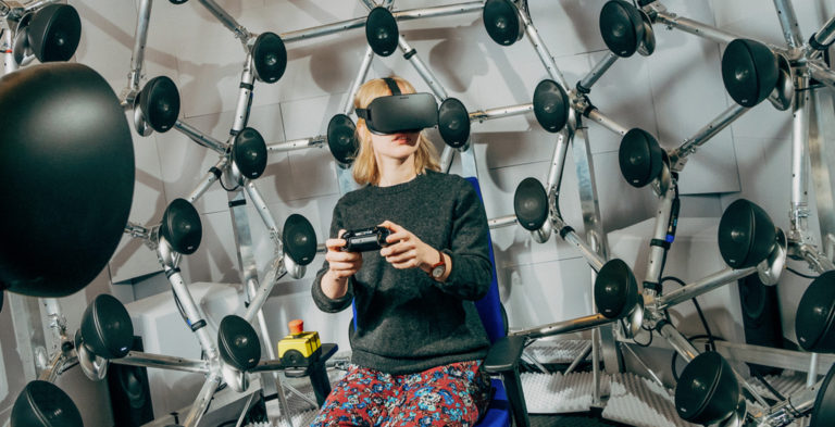 Lady sitting with a virtual headset on and holding a controller surrounded by speakers