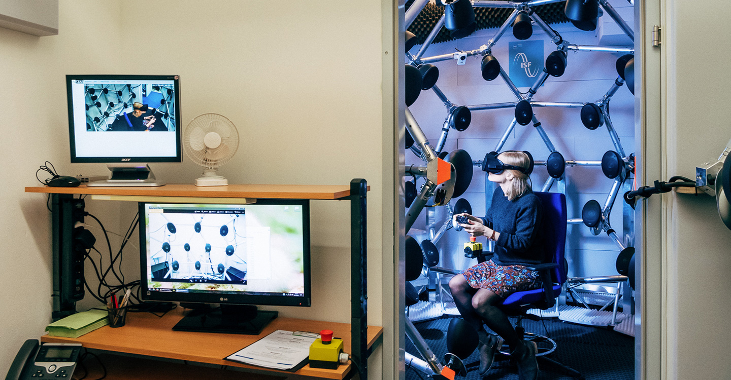 two rooms with computers in the first and a person in the second wearing a virtual headset and surrounded by speakers